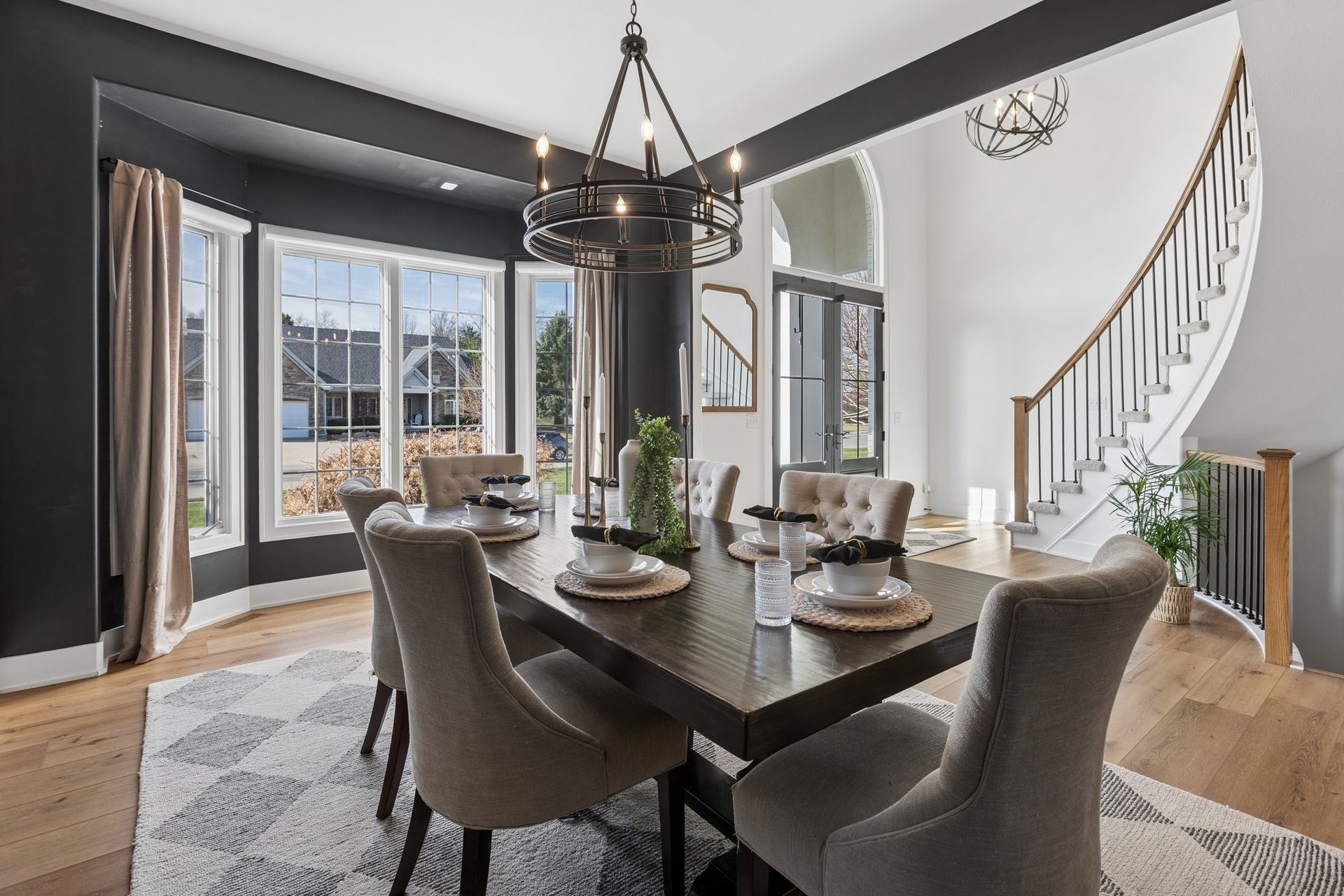 Open dining room with dark table, upholstered chairs, chandelier, and curved staircase beside large windows.