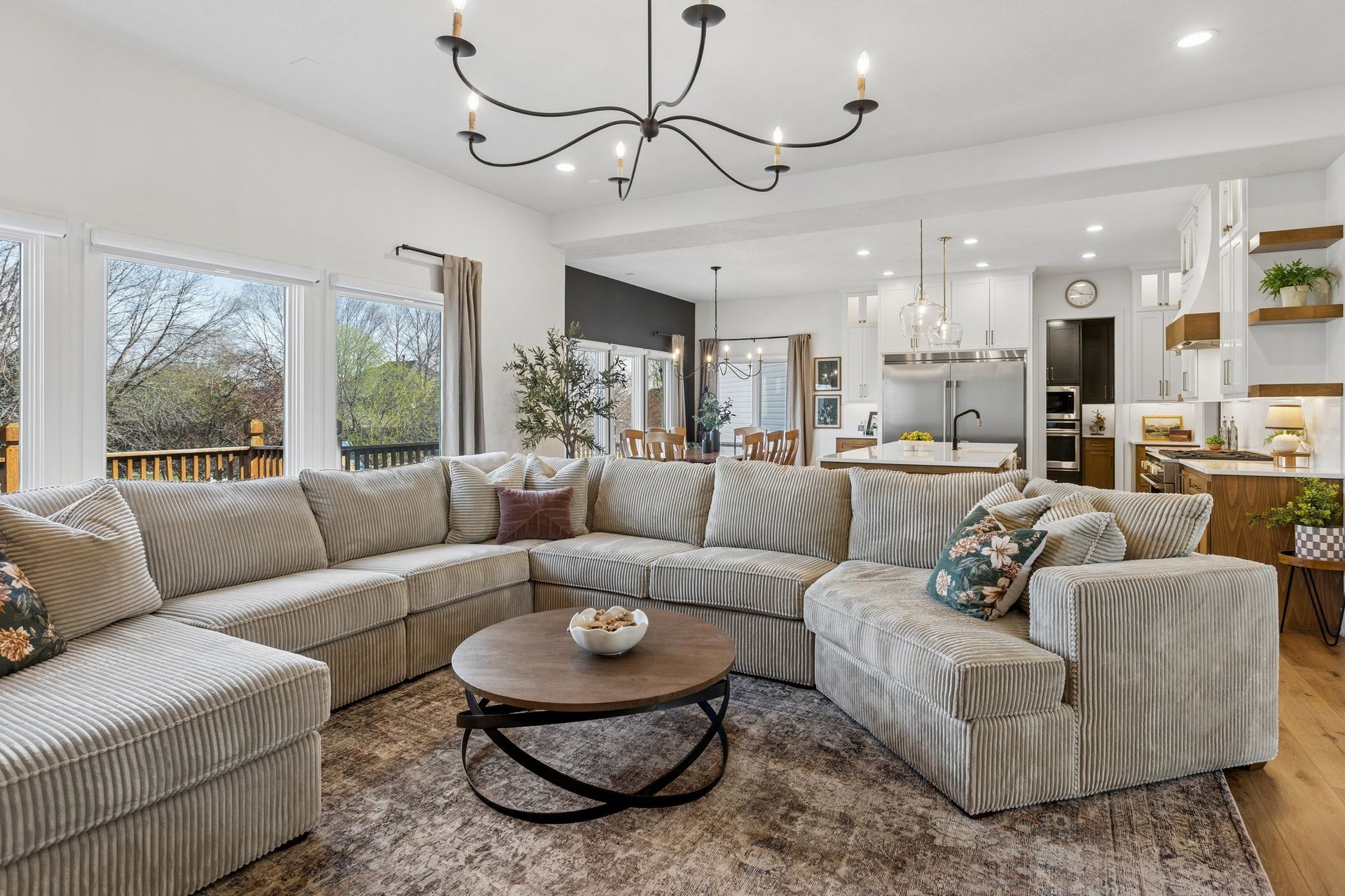 Bright open living room with a beige sectional, round coffee table, chandelier, and kitchen in the background