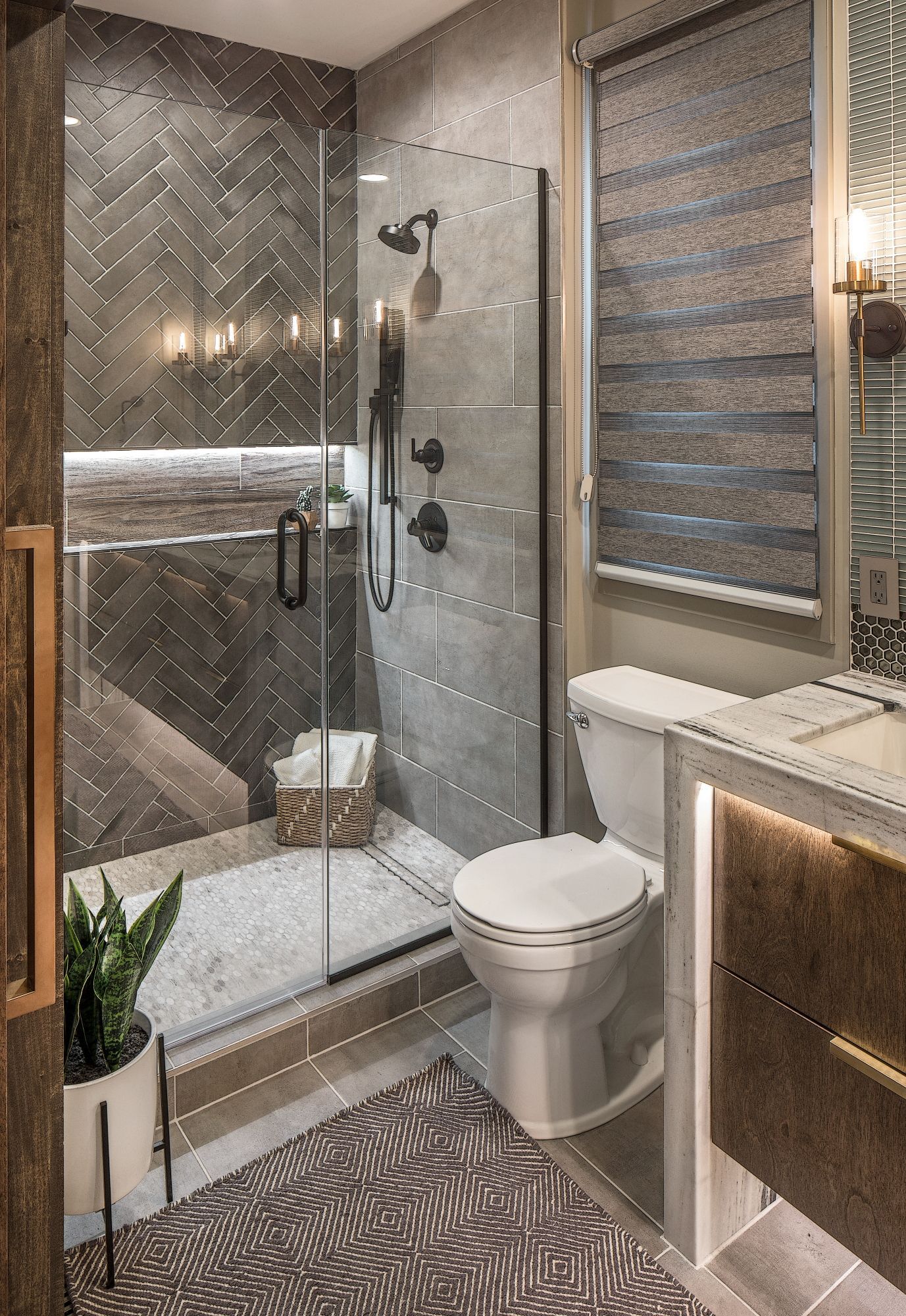 Compact luxury bathroom with glass shower, toilet, vanity, patterned tile, and woven blinds.