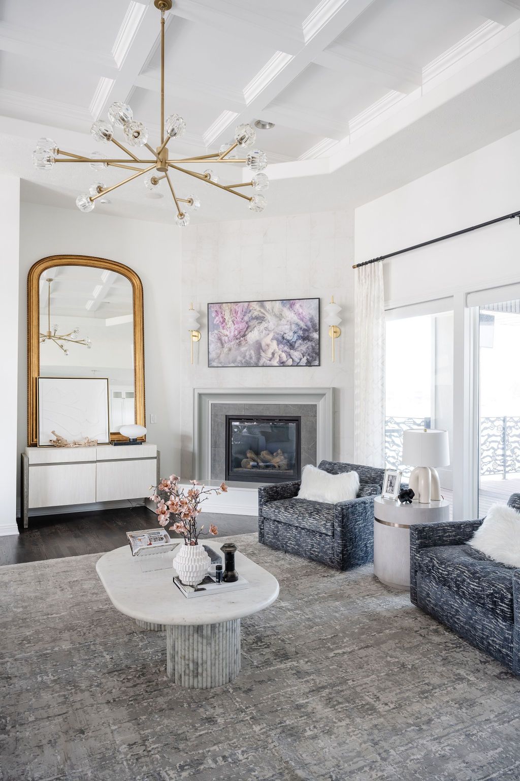 Bright, elegant living room with gray sofas, fireplace, gold mirror, and large chandelier