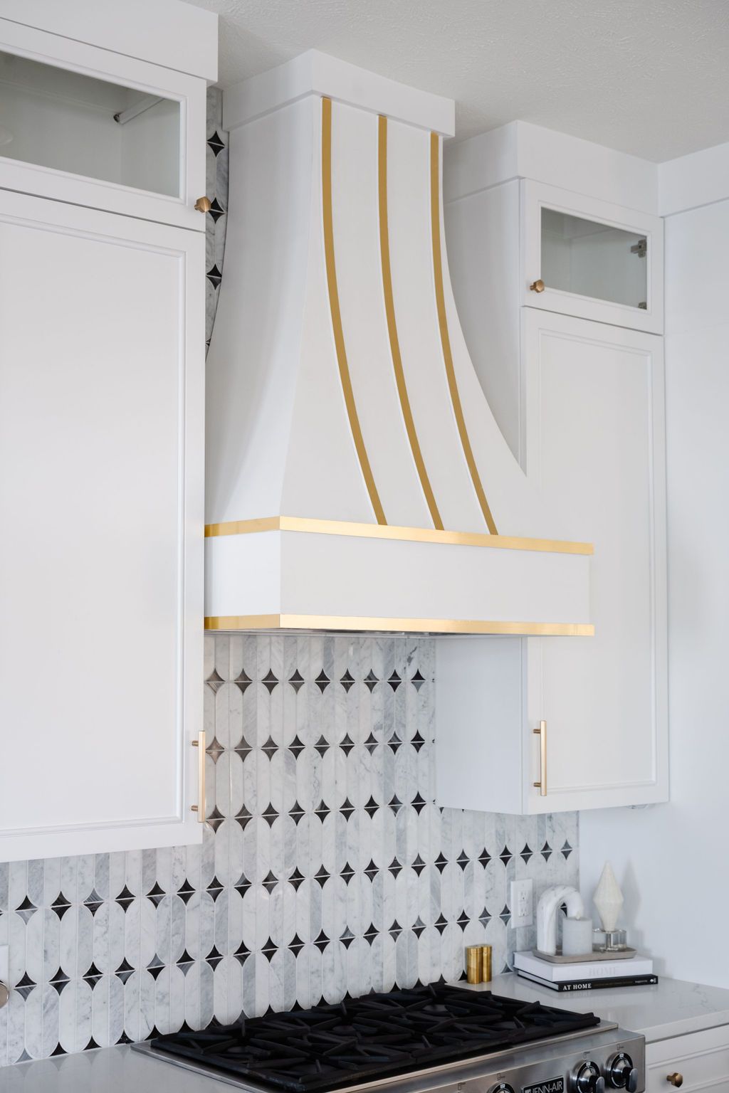 White kitchen with gold-accented range hood, patterned backsplash, and black stove tops