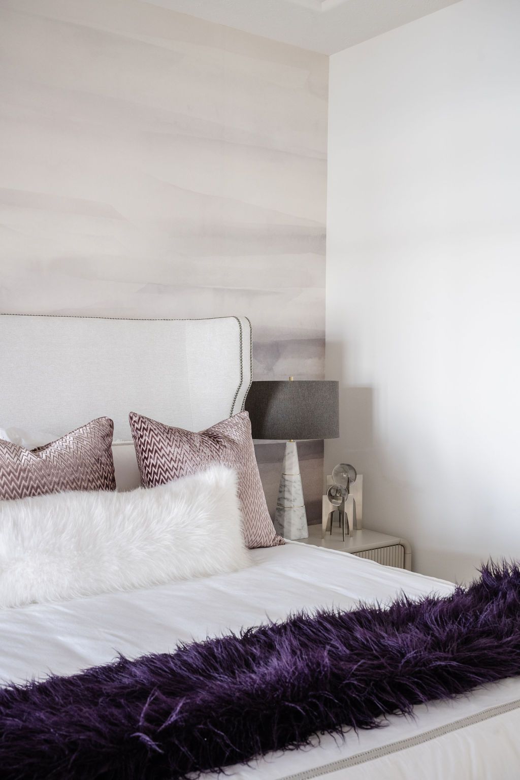 Bright bedroom corner with a white bed, patterned pillows, gray lamp, and purple throw blanket