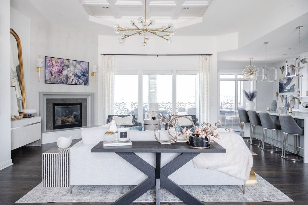 Bright white living room with fireplace, chandelier, large windows, and black coffee table with decor.