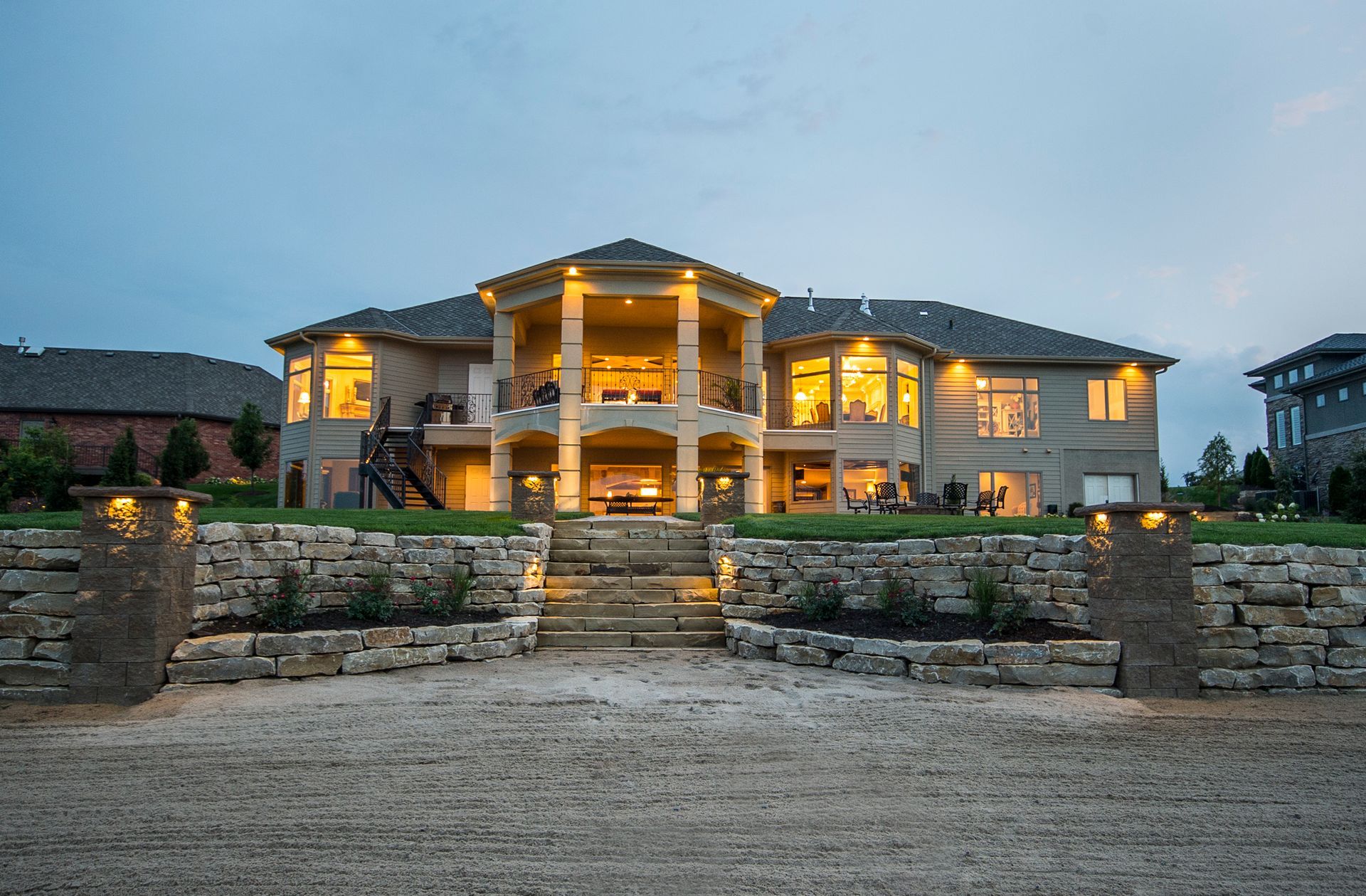 Large illuminated luxury house at dusk with stone terrace and landscaped entrance