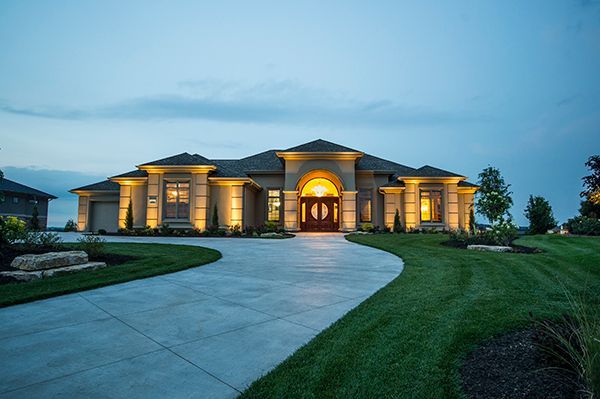 Large stone house at dusk with warm lit entrance, curved driveway, and manicured lawn