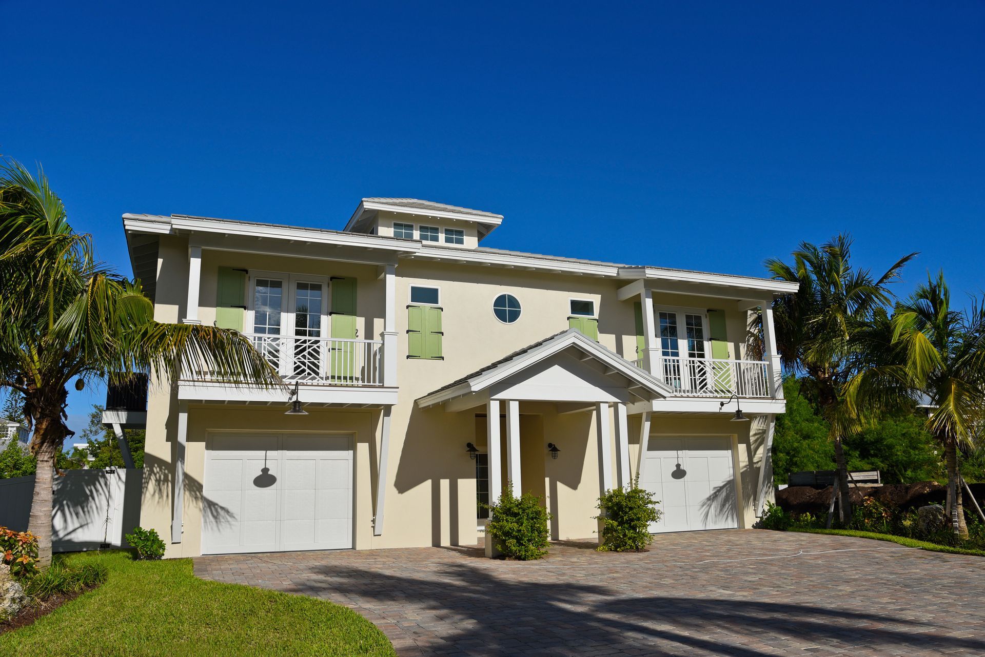 Two-story beige house with white trim, balcony, garage, and a palm-lined driveway under a clear blue sky