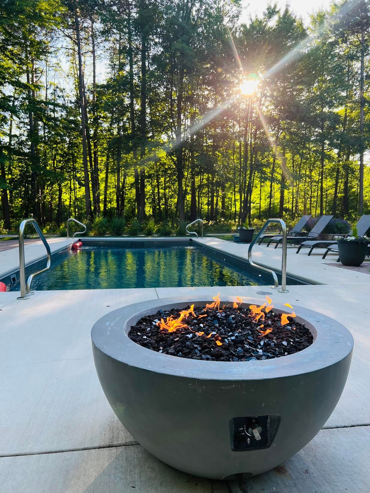 A fire pit in front of a swimming pool with the sun shining through the trees.