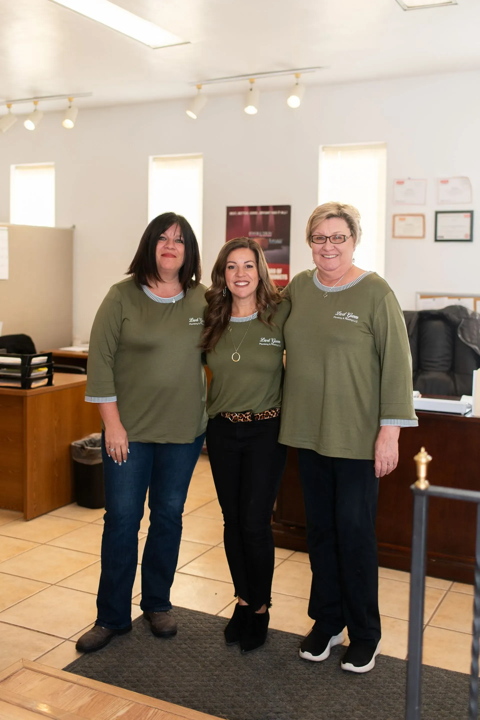 Three women standing in an office, wearing green shirts, smiling, the middle woman with arms around the other two.