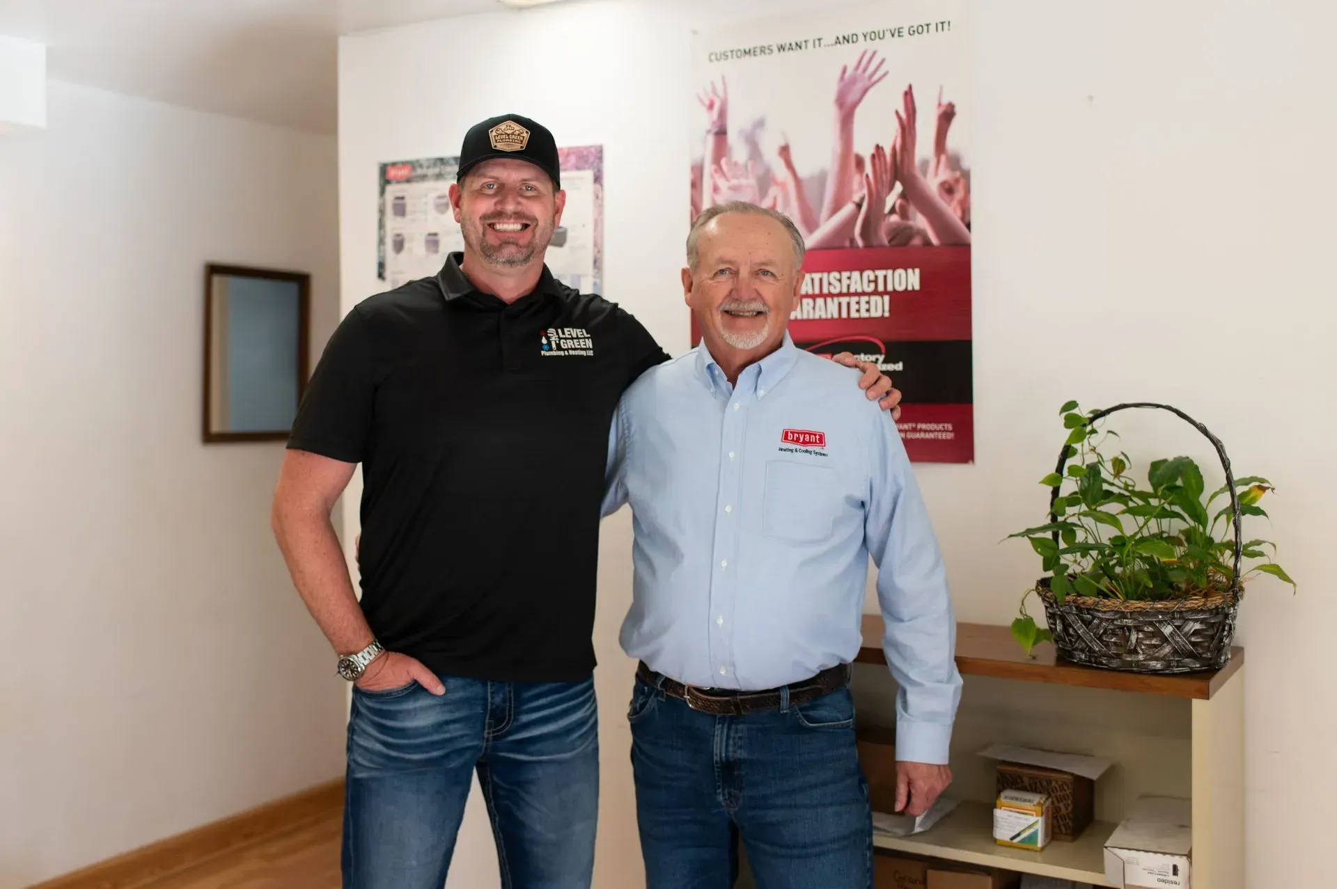 Two men standing next to each other, one in black shirt and jeans, the other in a blue shirt and jeans, smiling in an office.