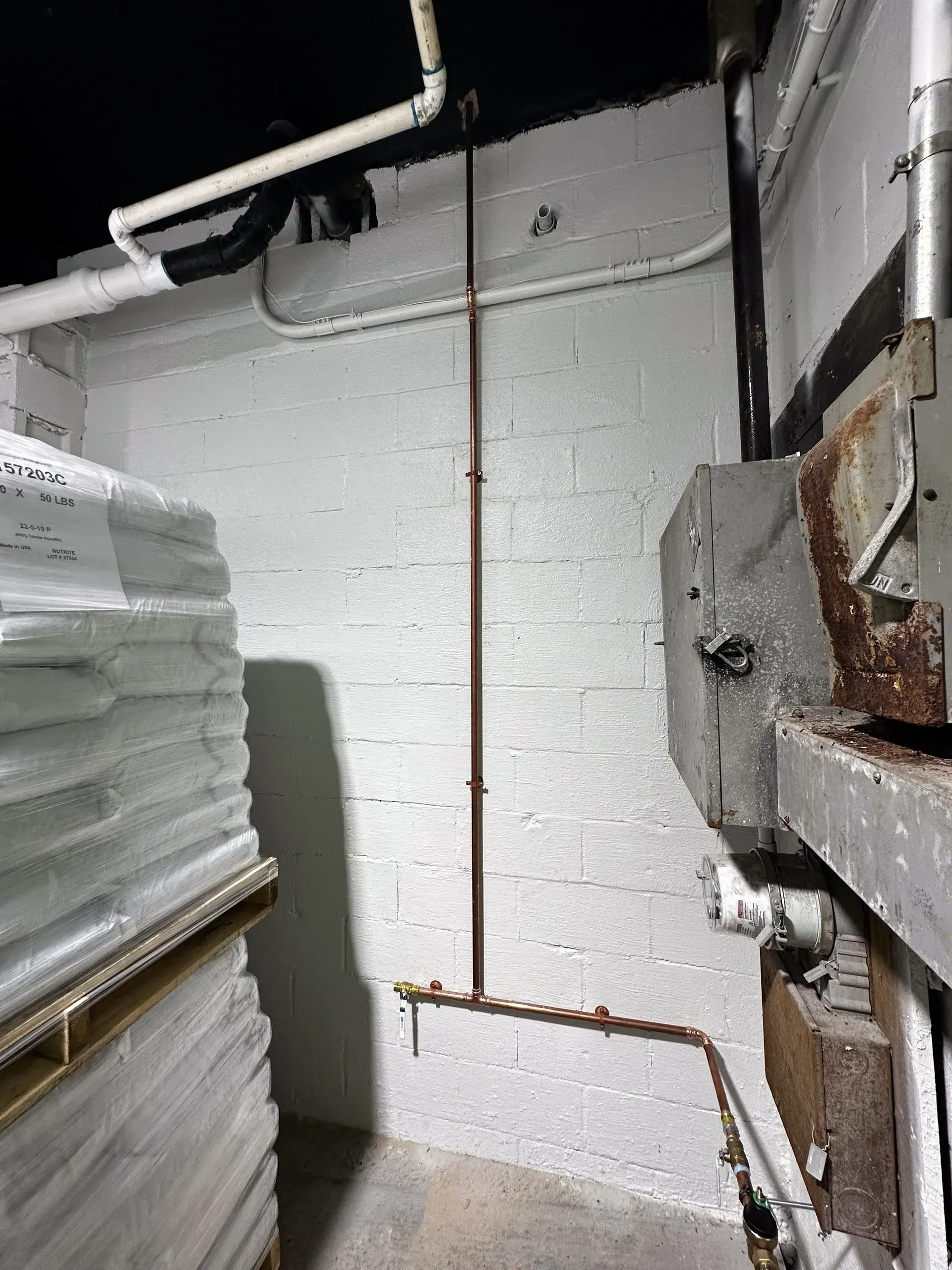 Copper pipe running up a white brick wall in a basement; a stack of white foam sheets is on the left.