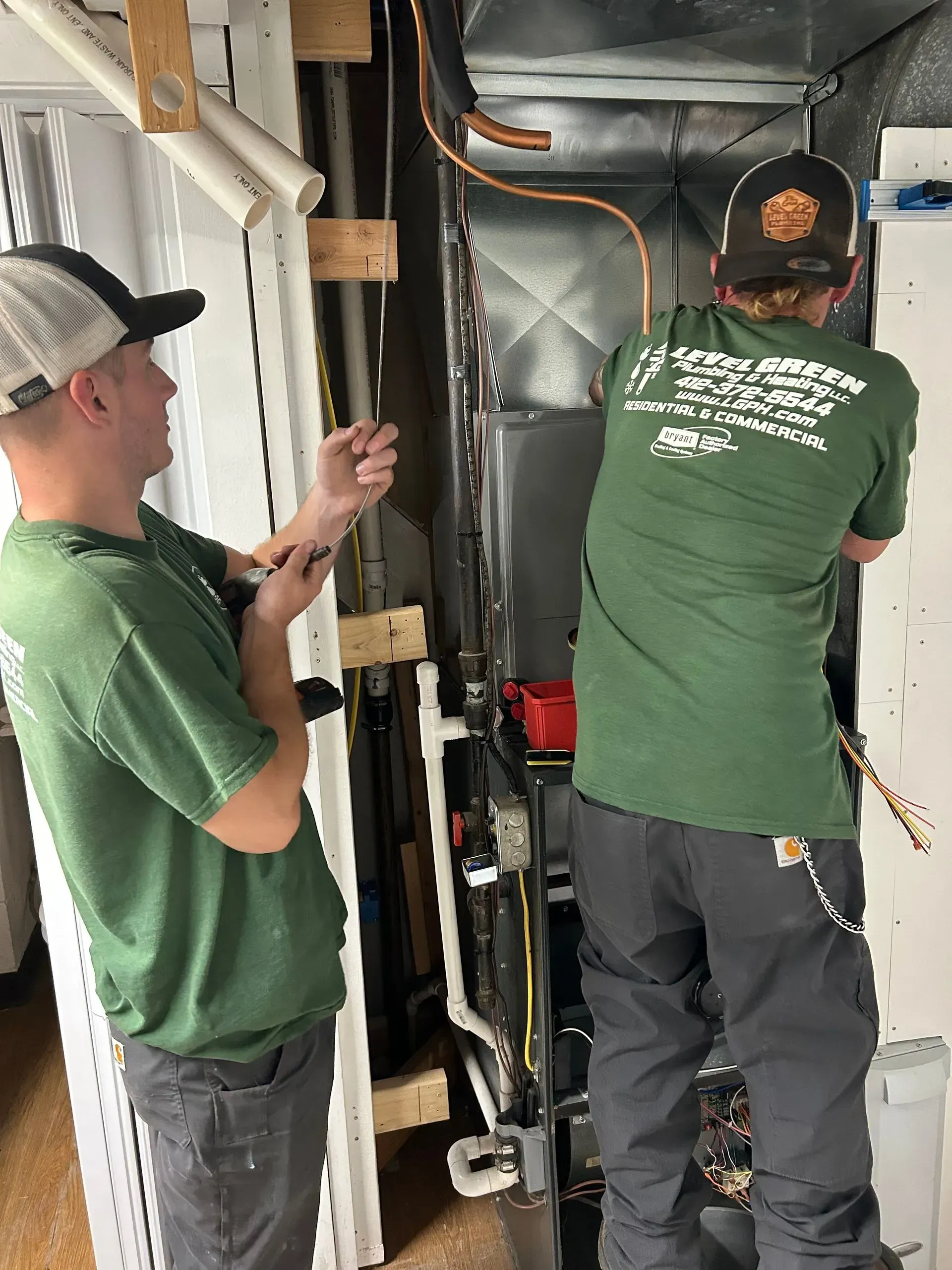 Two HVAC technicians working on an air handler unit. One holds a wire, the other works on copper tubing.