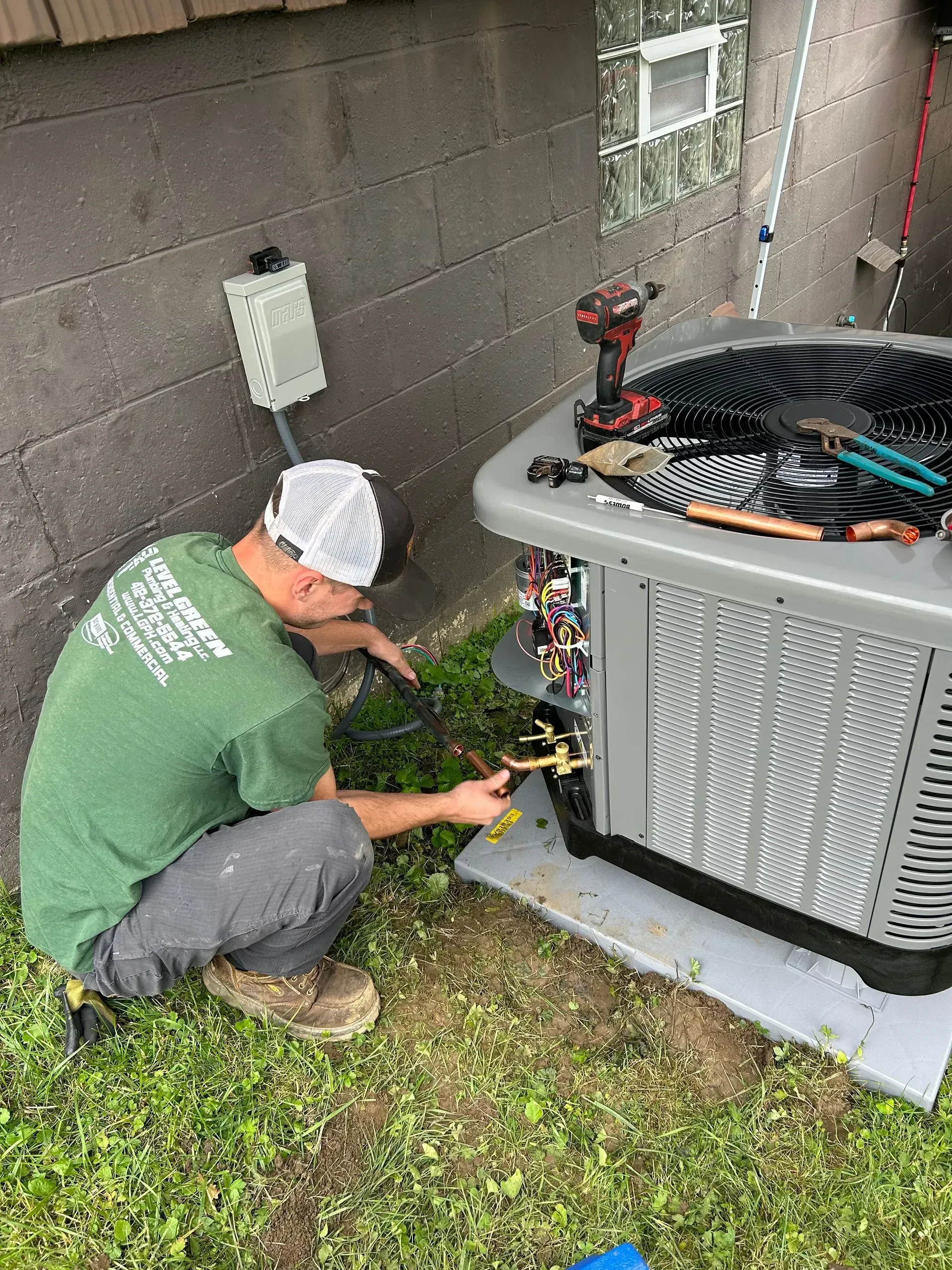 HVAC technician in green shirt repairing an air conditioner unit next to a brick building.