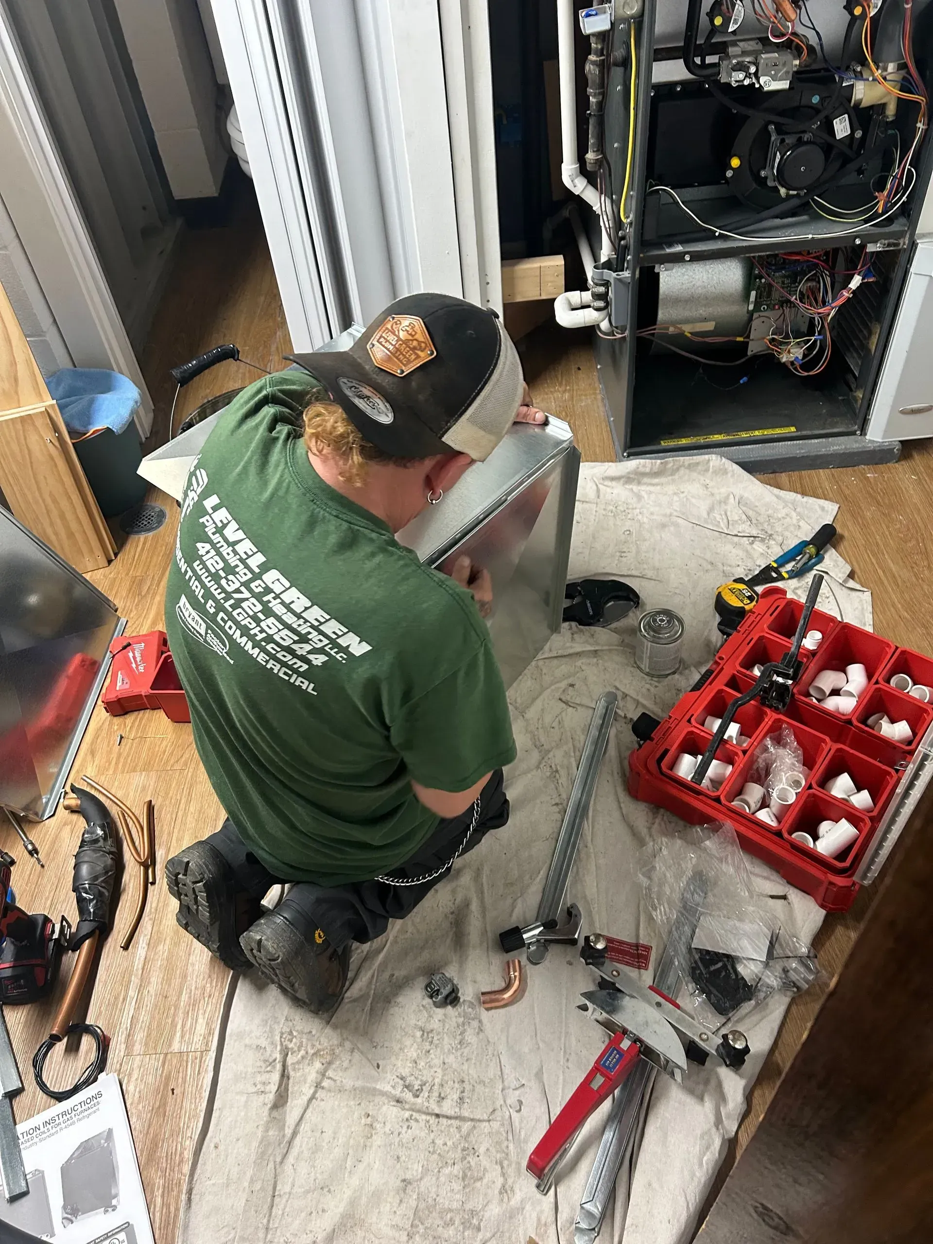 HVAC technician kneels, working on furnace. Wearing a green shirt, he is surrounded by tools.