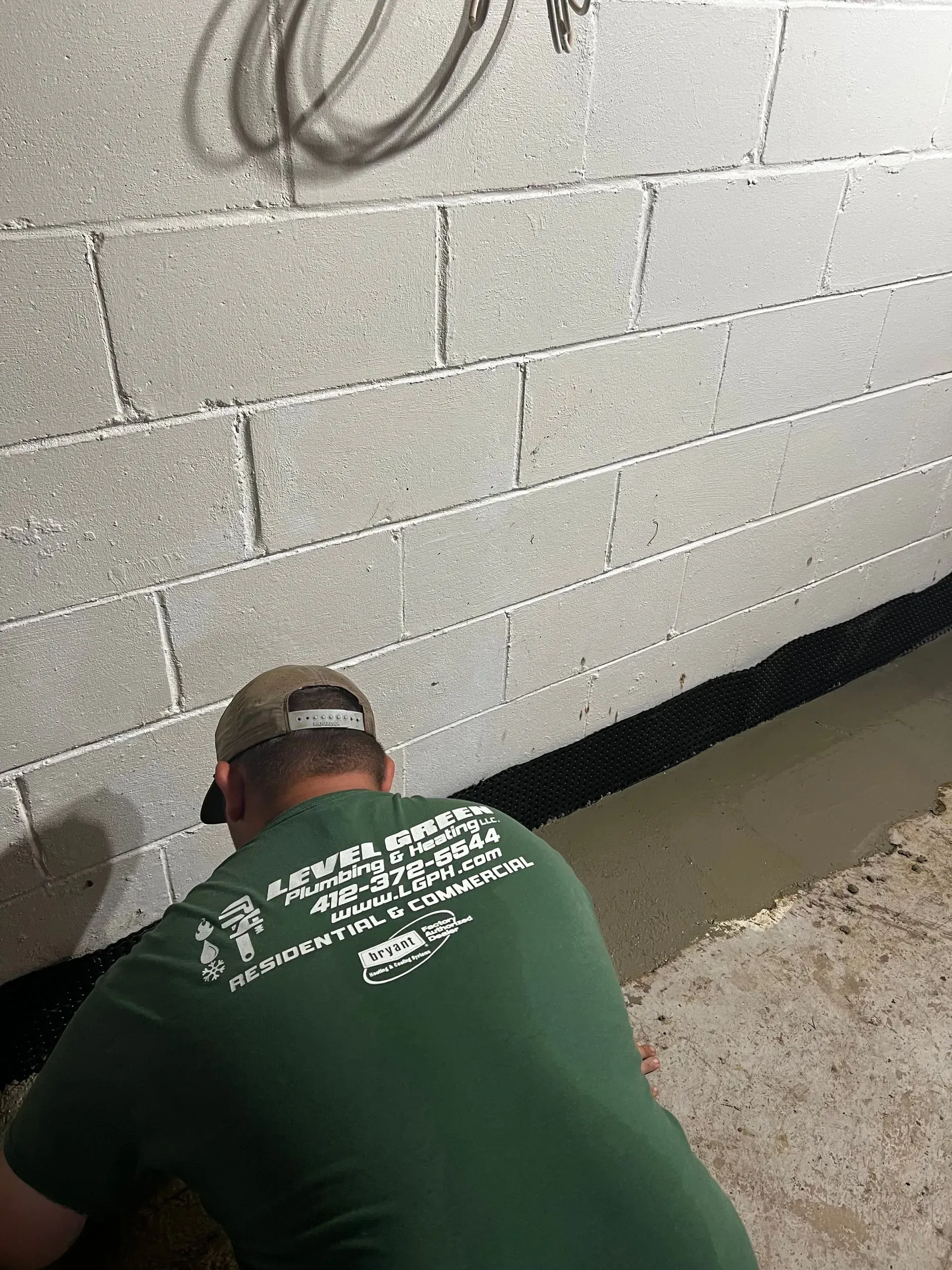 Man in green shirt installing waterproofing along a cinder block basement wall.