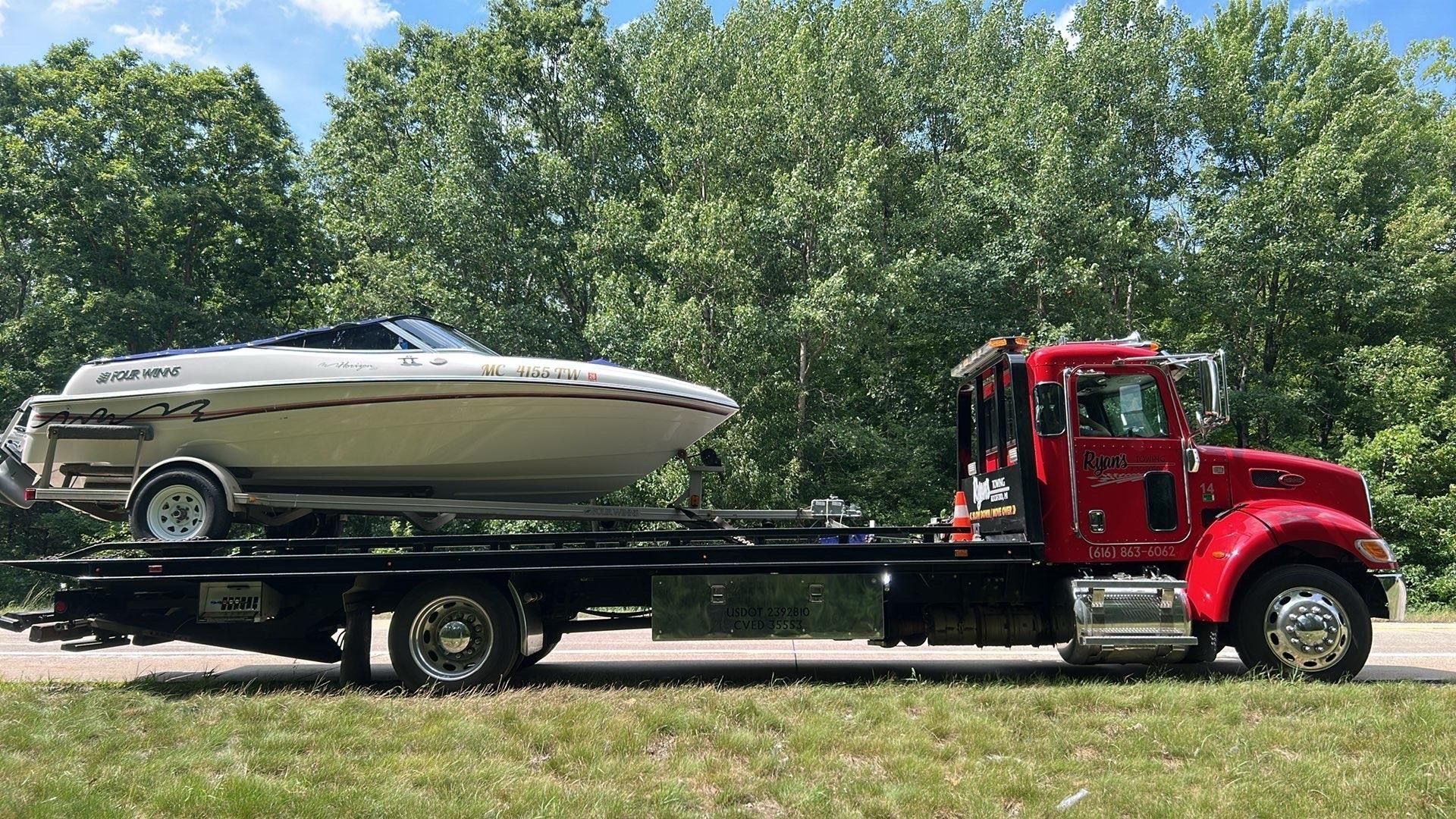 Red tow truck carrying a white boat on a trailer, next to green trees on a sunny day.