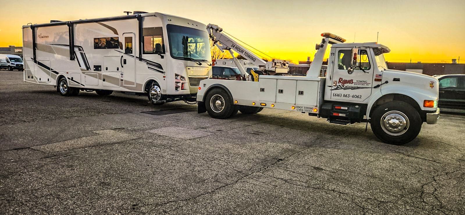 A white tow truck towing a white RV on a gravel lot at dusk.