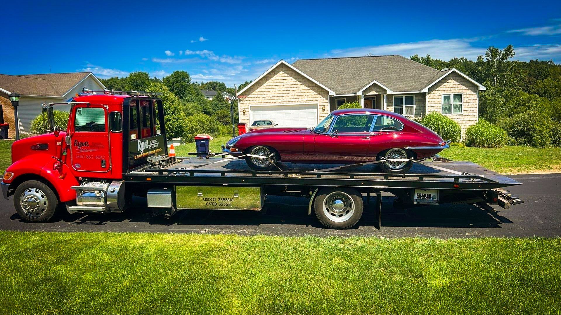 Red tow truck transporting a maroon Jaguar sports car in front of a house on a sunny day.