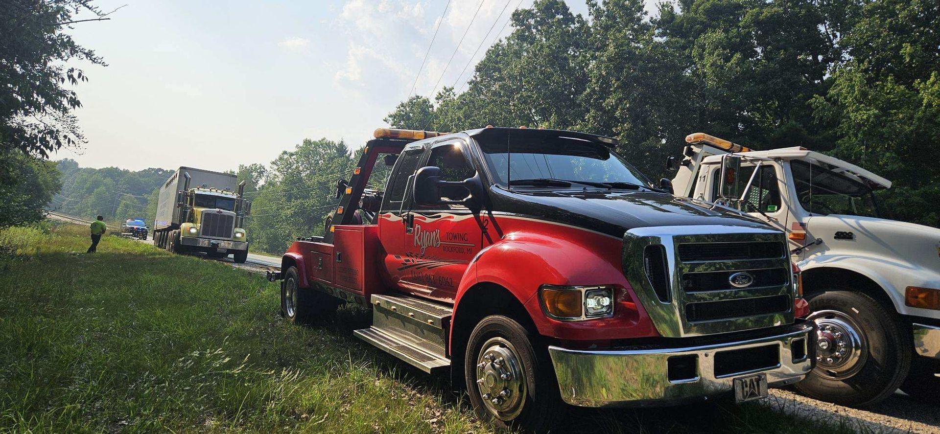 Red tow truck and white tow truck next to a road, where a semi-truck is stopped. Trees in background.