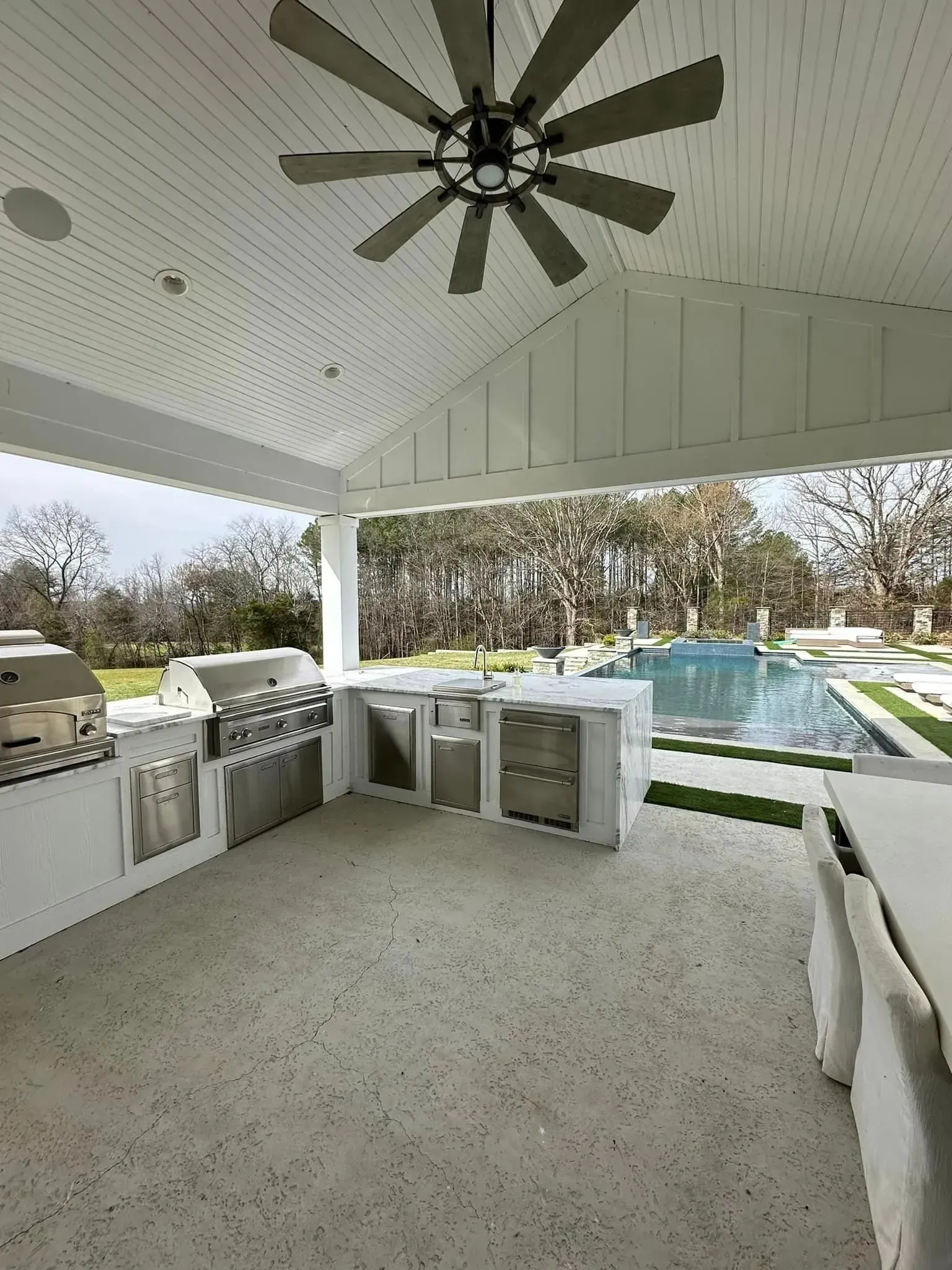 Outdoor kitchen with grills, refrigerators, and a ceiling fan, overlooking a pool.
