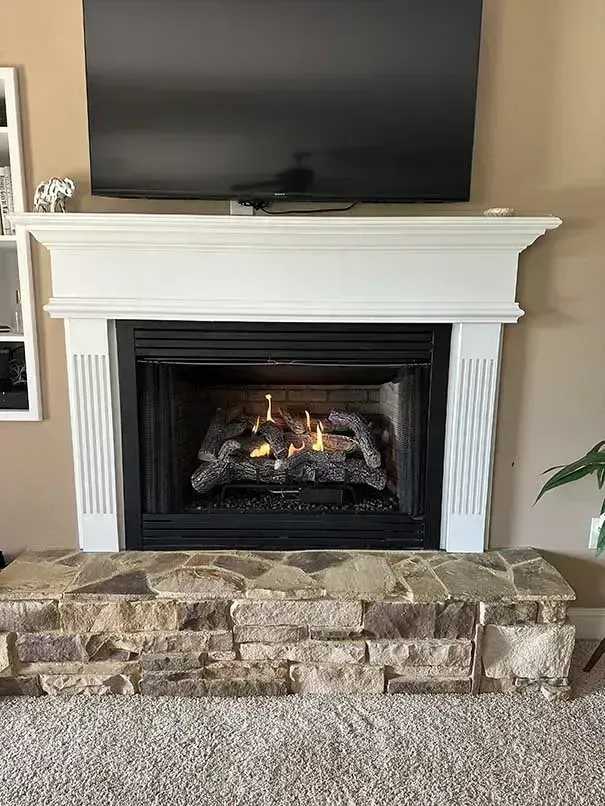 Fireplace with stone base, white mantle, and lit gas logs. Television mounted above.