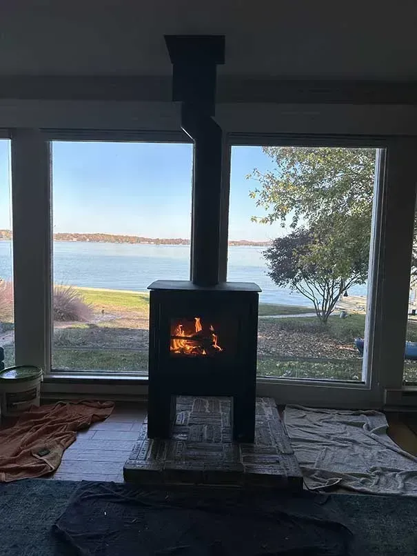 Wood-burning stove with flames, chimney, and a waterside view through a large window.