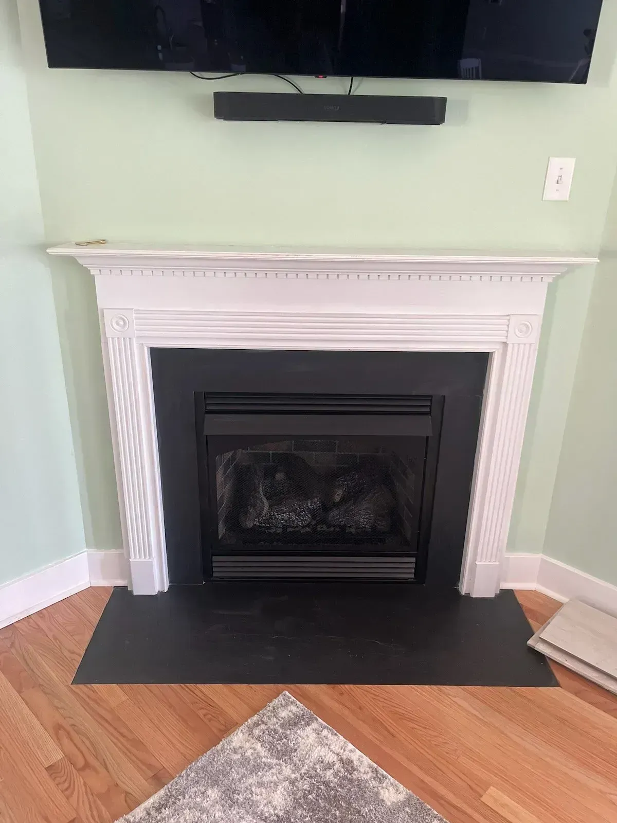 Fireplace with white mantel, black firebox, and TV above, set in a corner of a room with light green walls and wood floor.