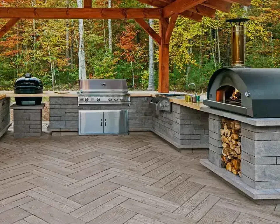 Outdoor kitchen with grill, pizza oven, and wooden pergola. Wood and brick construction on a patio.