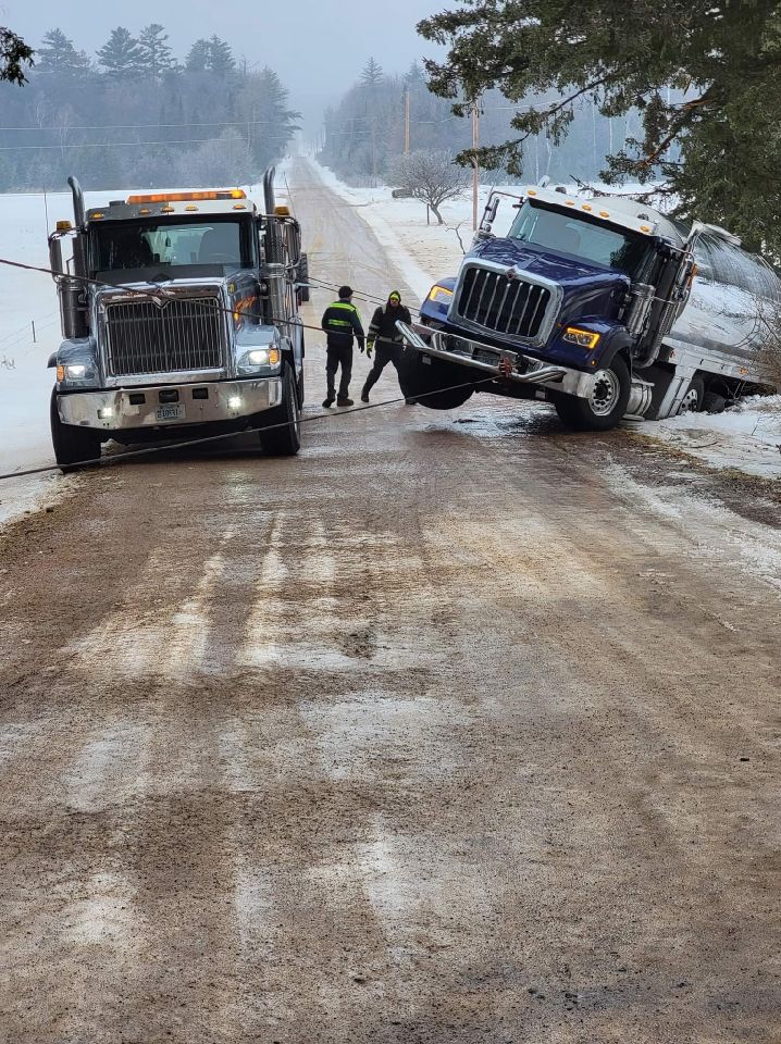 Tow trucks and workers at a snowy roadside crash, with a blue semi-truck tilted off the road.