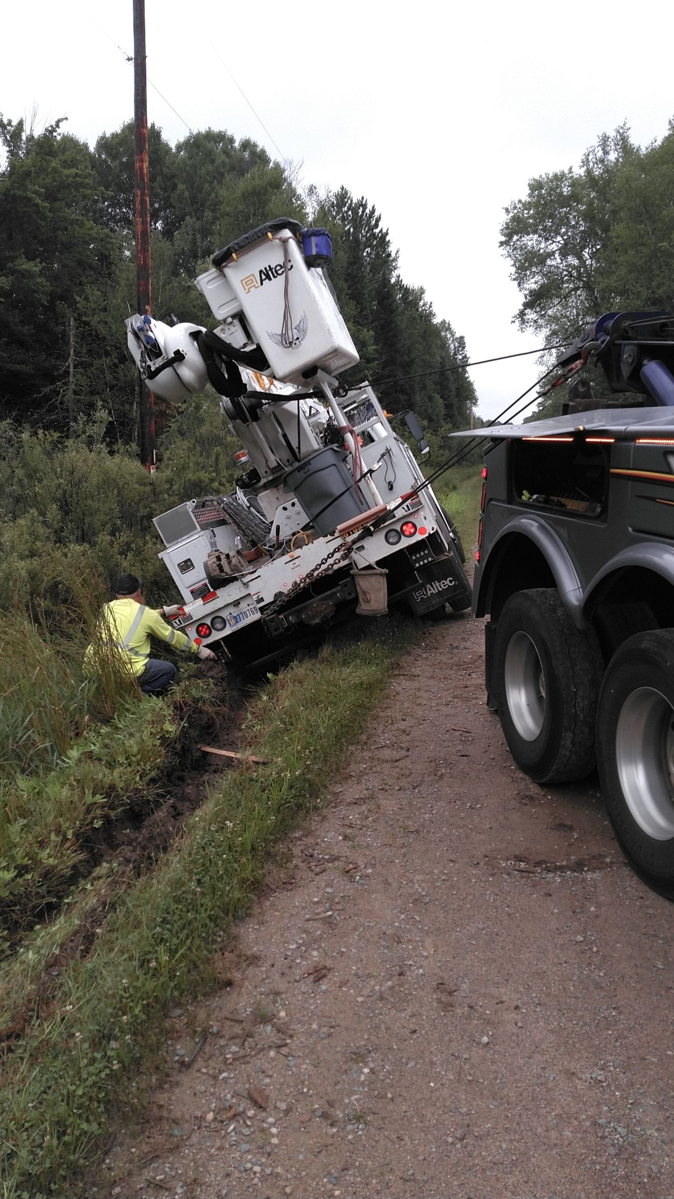 Overturned utility truck in a ditch beside a dirt road, with a tow truck nearby.