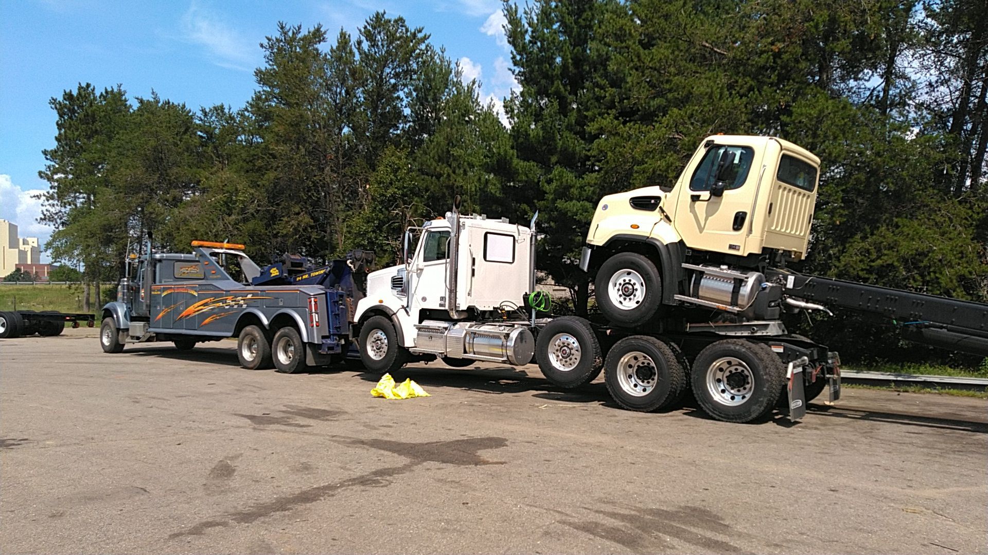 Tow truck hauling a yellow vehicle on a flatbed in a parking lot, with trees in the background