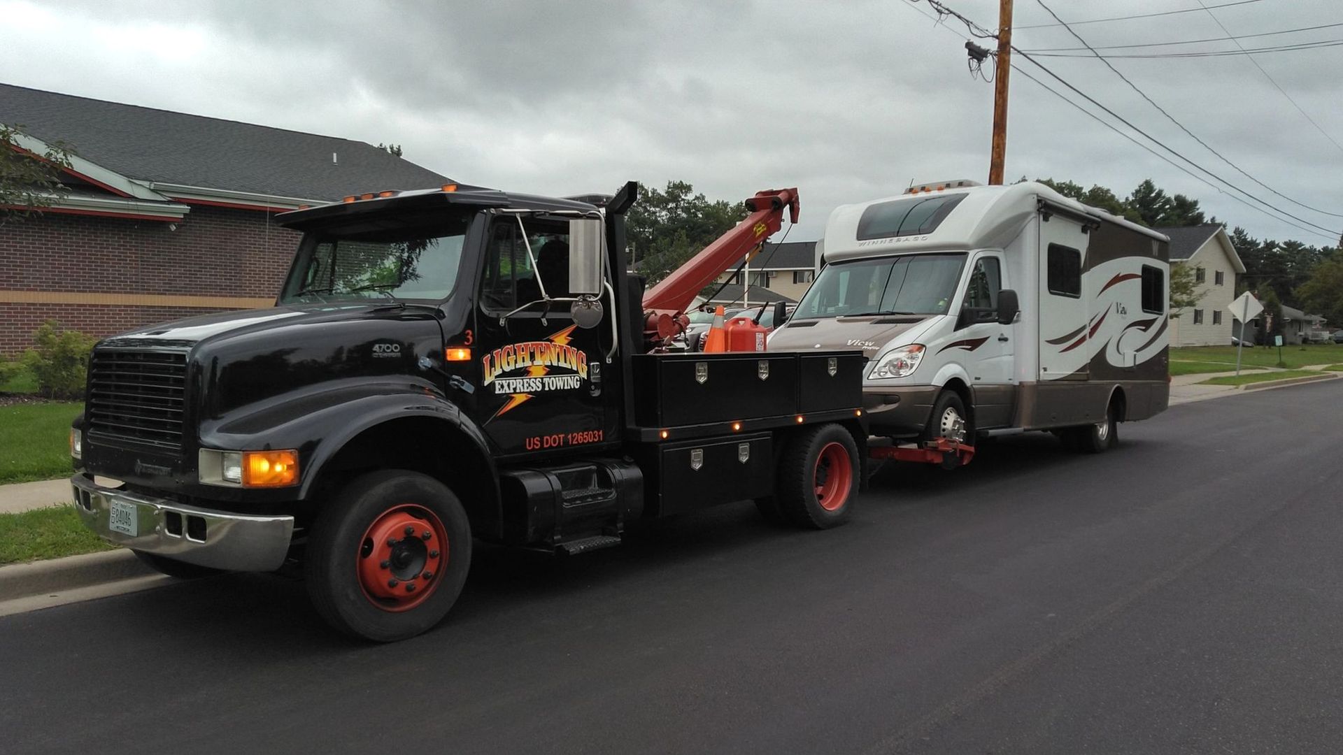 Tow truck hauling a white RV on a suburban road under cloudy skies