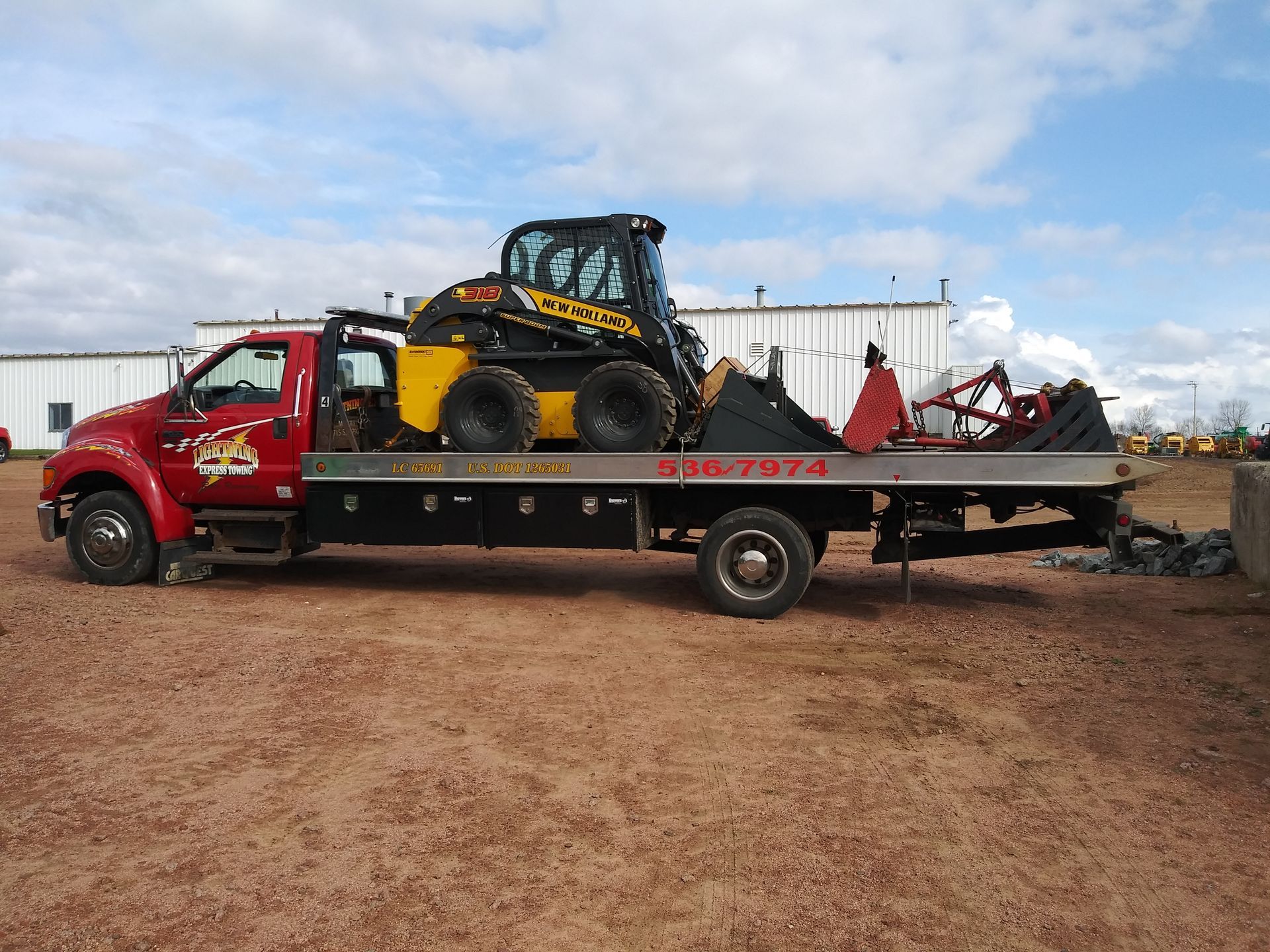 Red flatbed tow truck carrying a yellow skid steer and equipment on a dirt lot under a cloudy sky