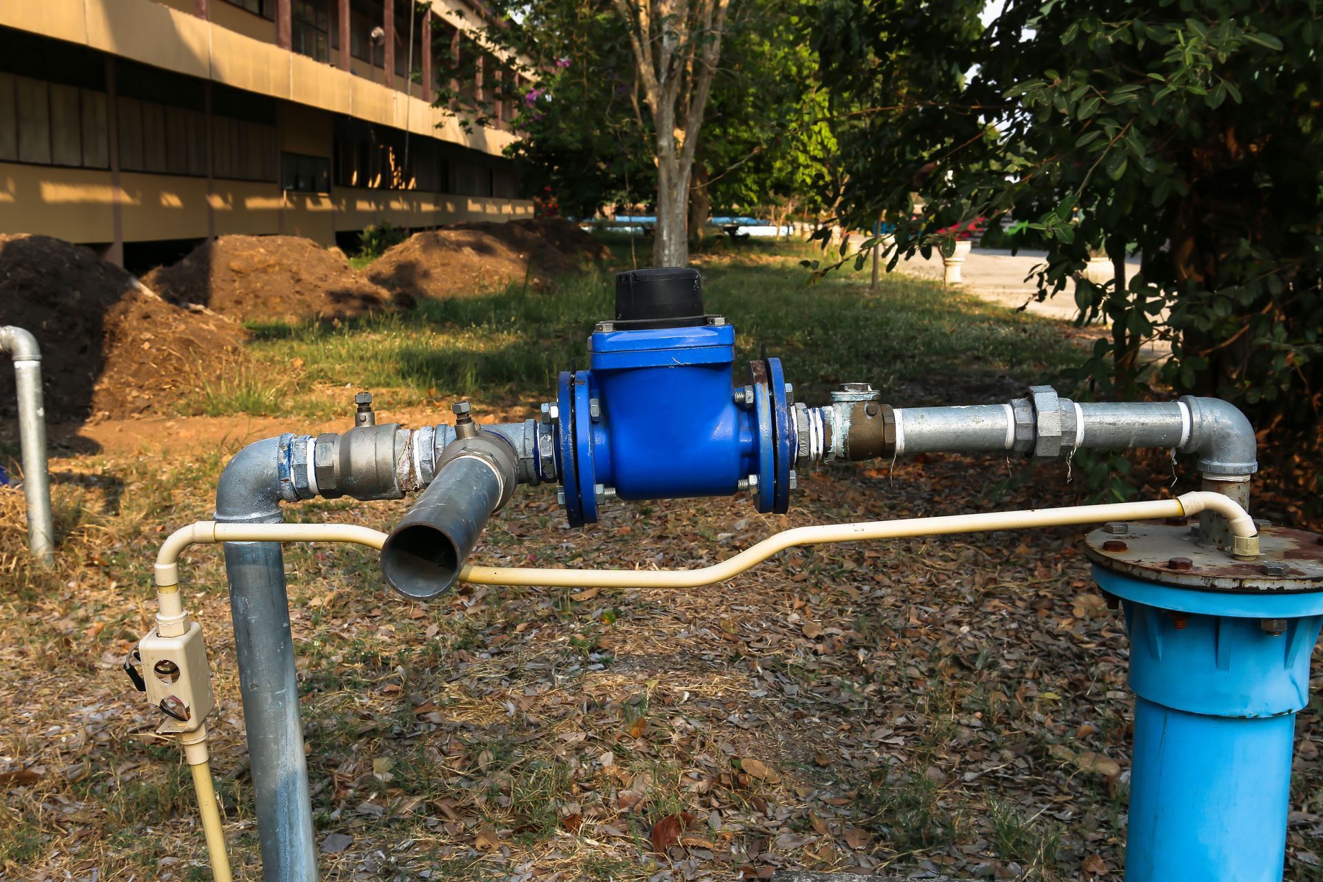 Blue industrial pipe assembly with valves and a large horizontal pipe outdoors under trees