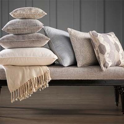 A bench with a stack of neutral-toned pillows and a fringed blanket; against a gray wall.