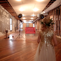 Woman in gold-sequined gown stands in a large, unfinished room, facing a red bench.