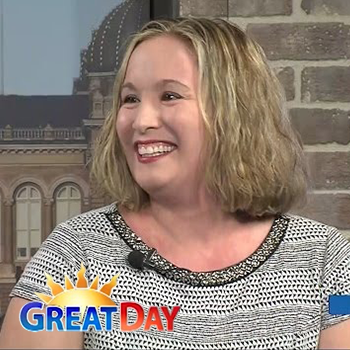 Woman with blonde hair smiles, wearing a black and white patterned top, sitting on a set with a 