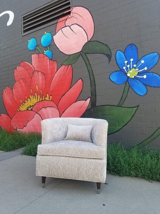 Chair in front of a flower mural on a gray brick wall; sidewalk in foreground, grass on either side.
