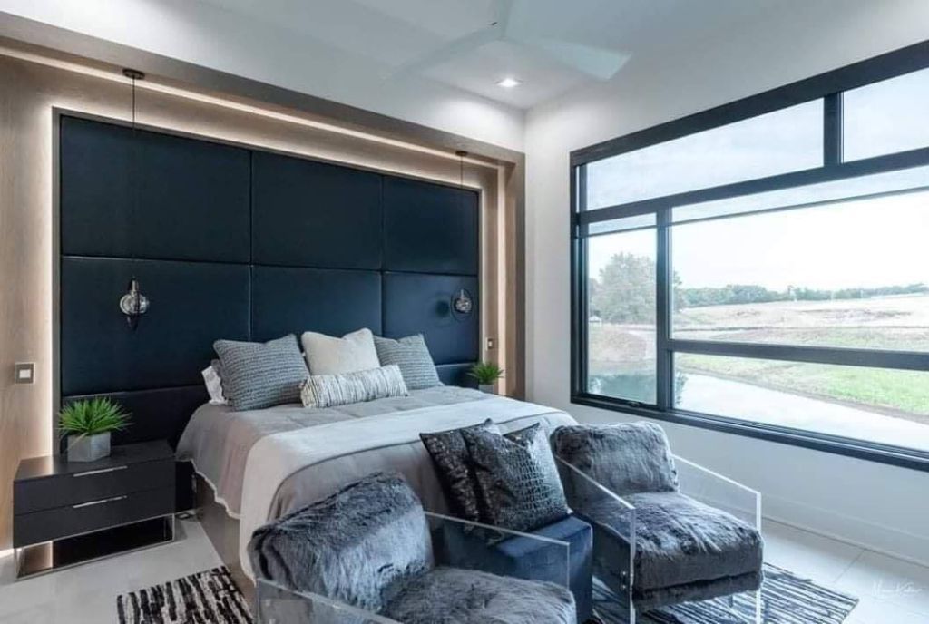 Modern bedroom with a bed, two chairs, large window, and dark blue headboard with ambient lighting.
