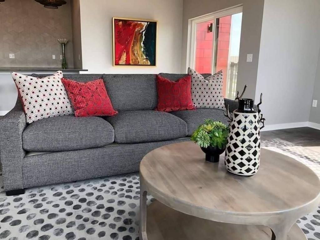 Living room with gray sofa, red pillows, art, round wood coffee table, patterned rug.