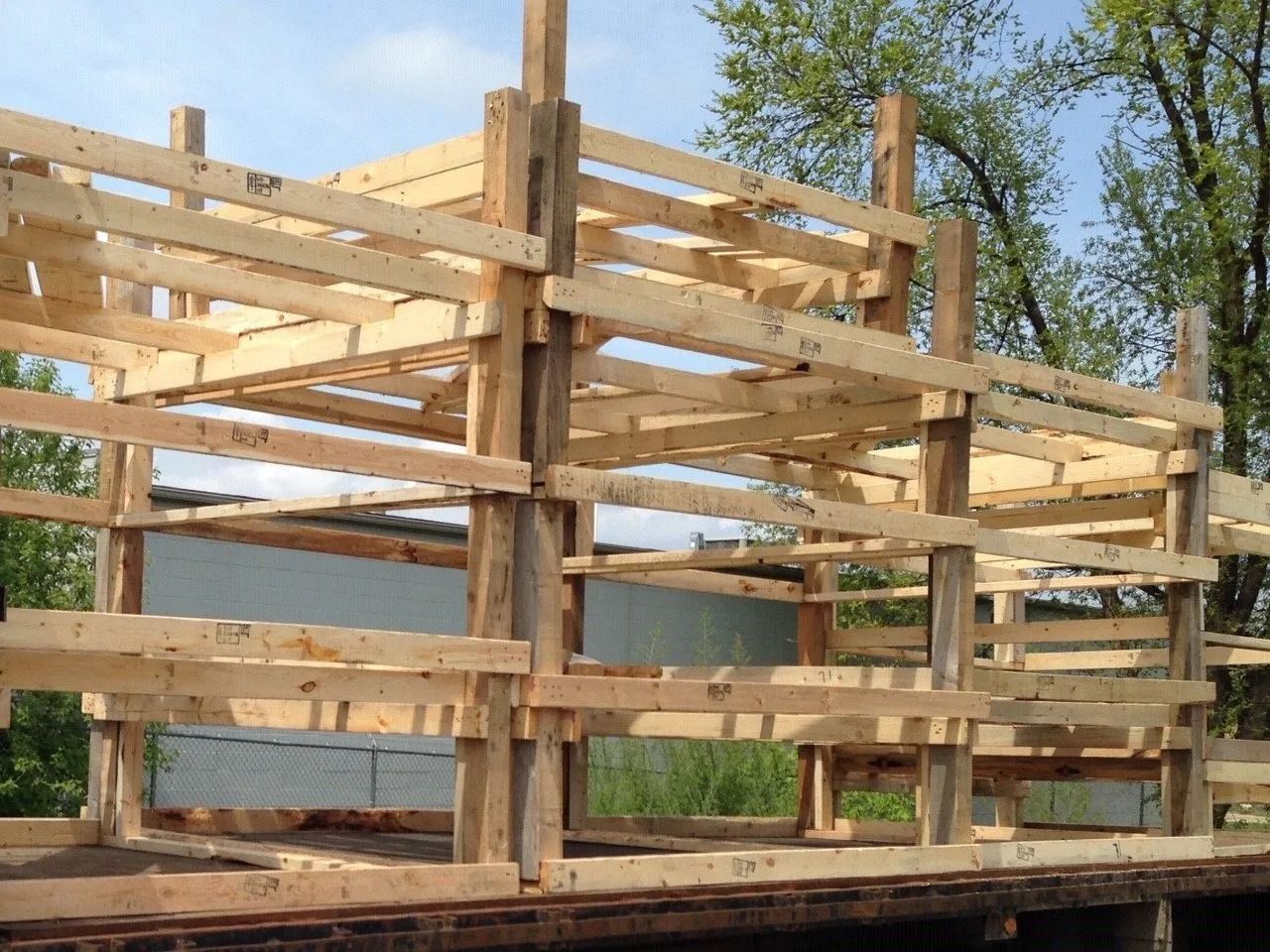 Wooden pallet structure under construction outdoors with horizontal slats and posts against trees and sky