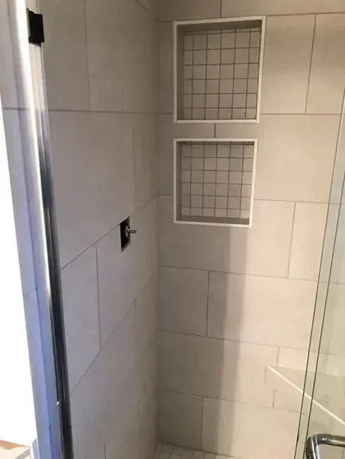 Shower interior with light gray rectangular tiles, two built-in niches with wire mesh, and glass door.