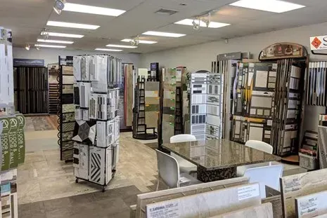Tile showroom interior with samples displayed on racks and tables, lit by overhead fluorescent lights.