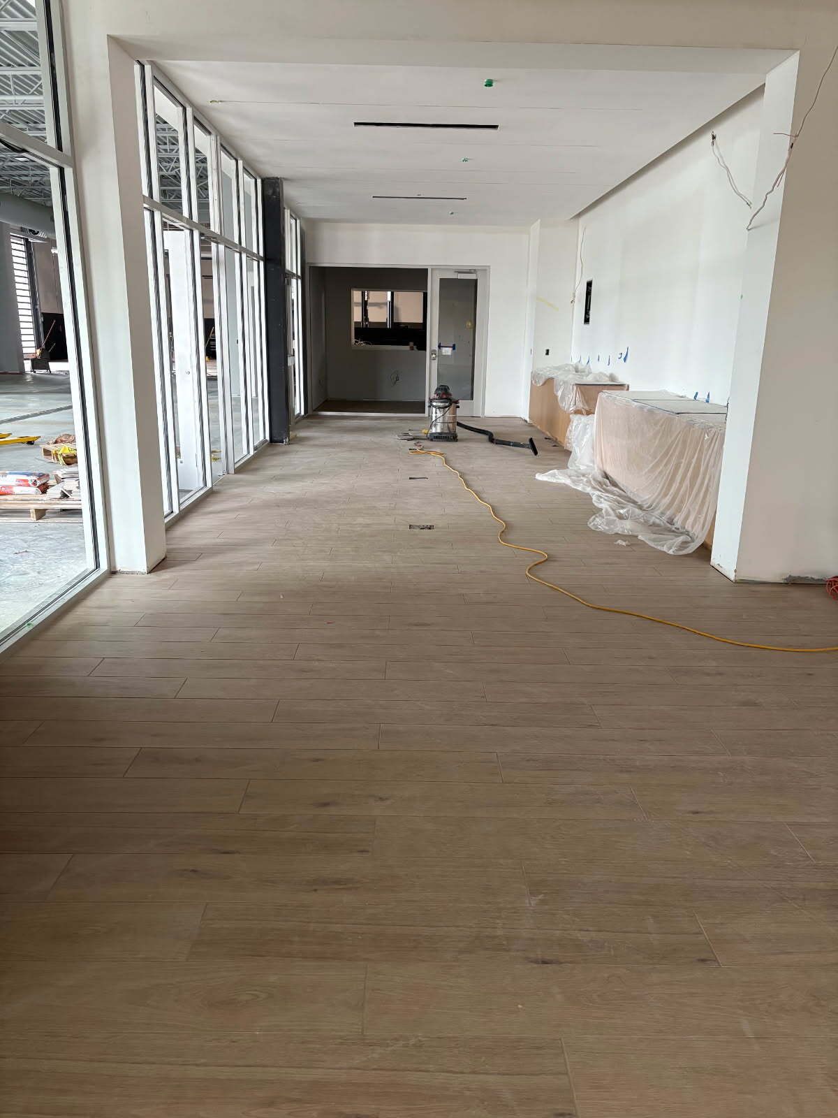 Interior hallway under construction with wood floors, floor-to-ceiling windows, and white walls.