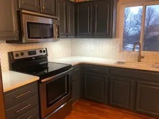 Gray kitchen cabinets with stainless steel appliances, white countertops and tile backsplash, wooden floors, and a window.