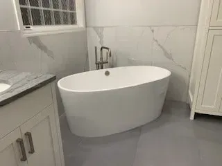 Oval bathtub in a modern white bathroom with marble-look wall tiles, cabinet, and gray floor.