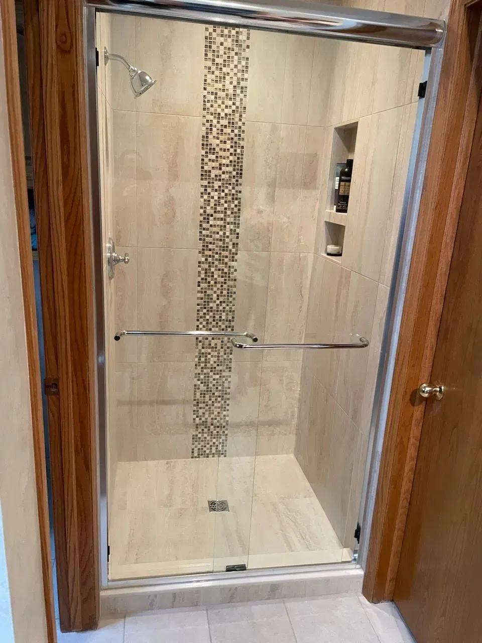 Walk-in shower with glass doors and tiled walls, featuring a mosaic strip and built-in shelves.