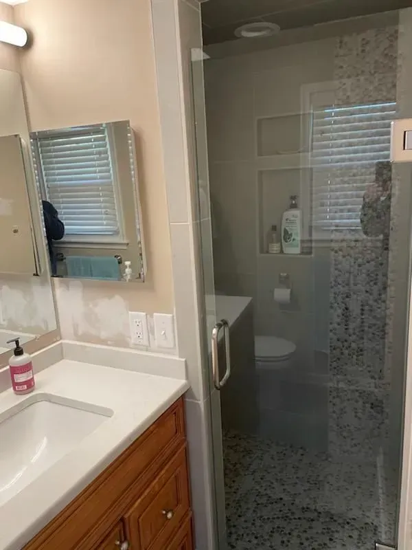 Bathroom with a sink, mirror, shower, and toilet. White countertops, wooden cabinets, and a glass shower door.