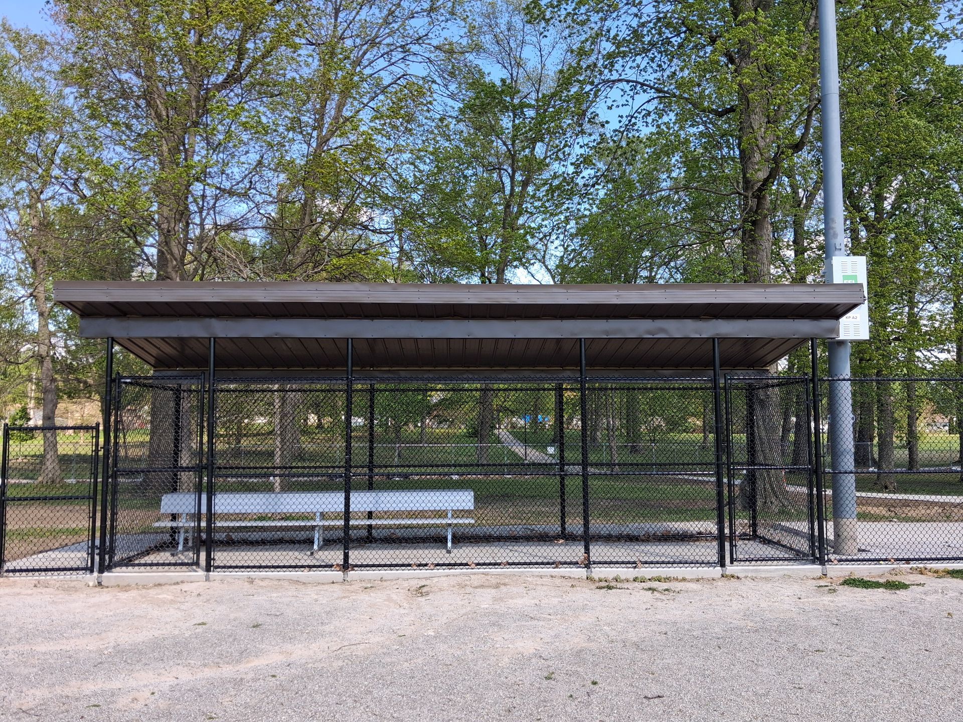 Covered outdoor bus stop shelter with benches, surrounded by trees and a chain-link fence.