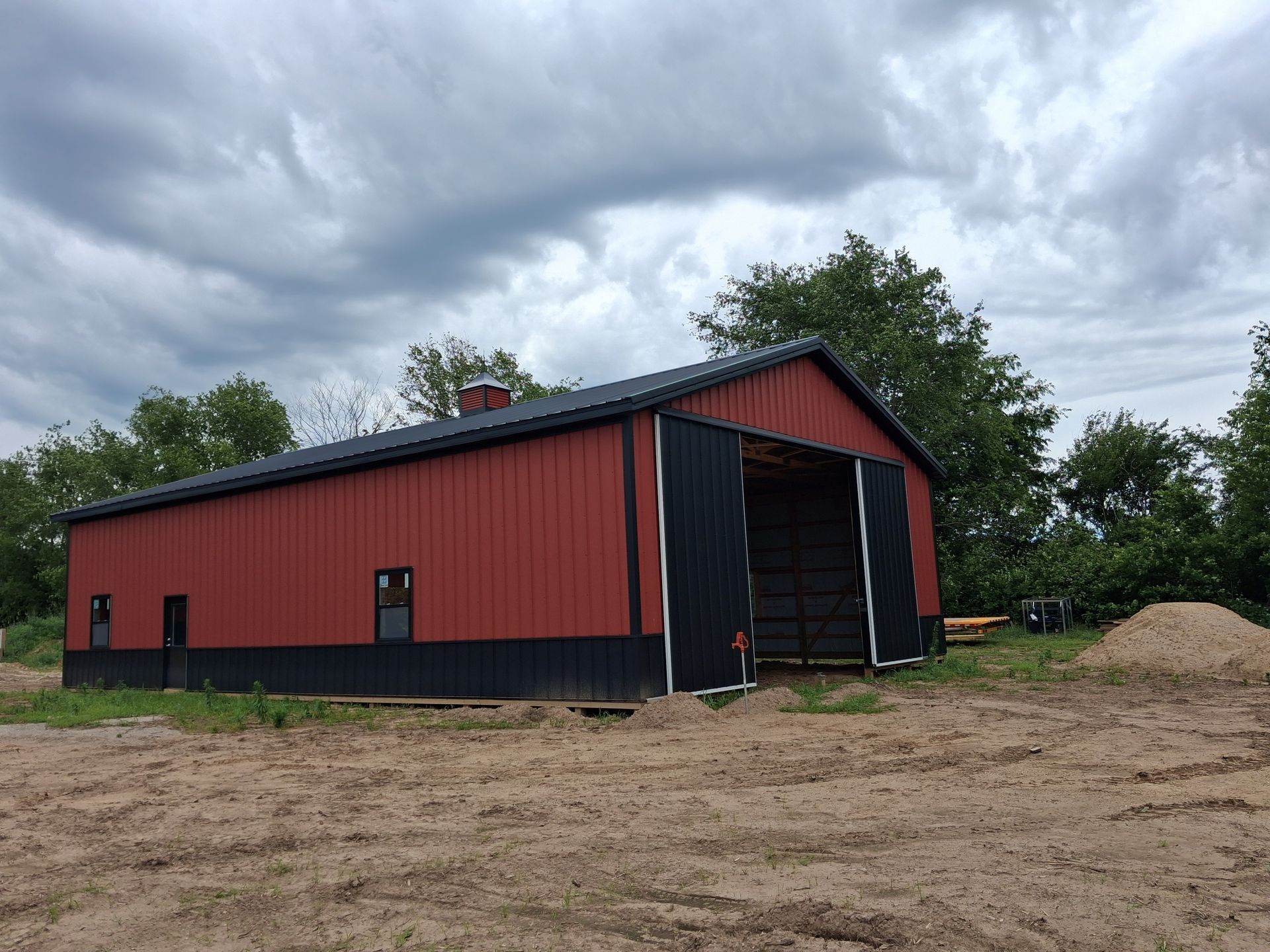 Red barn-style shed on a dirt lot under cloudy skies, with an open front and trees behind.