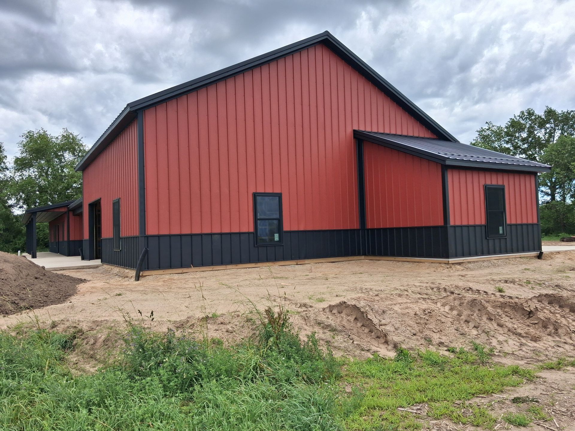 Red barn-style building with black trim on a dirt lot under cloudy skies